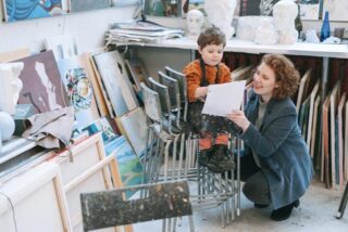 A female teacher holds a boy’s drawing in front of him surrounded by easels and art materials.