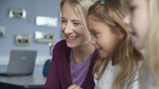 A female teacher and two pupils learning at a desk facing a laptop screen.