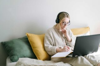 A woman taking notes whilst sitting on a bed with a laptop with headphones on.