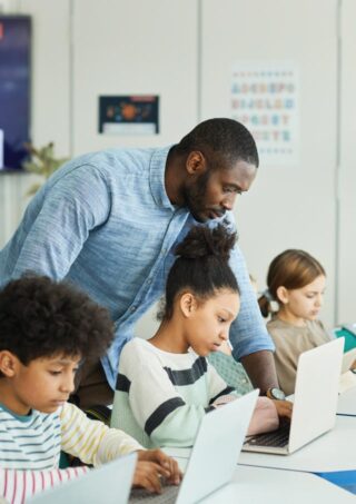 A male teacher assists a group of students working on laptops in a classroom.