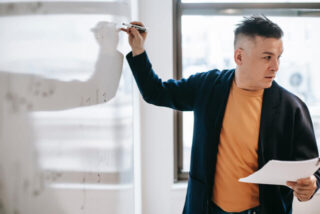 A male teacher holding a sheet of notes writes on a whiteboard.